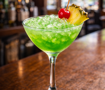 A green eyes cocktail recipe served in a cocktail glass with crushed ice, pineapple garnish, and cherry on a neighborhood bar counter.