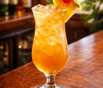 A jamaican iced tea cocktail recipe served in a hurricane glass with pineapple wedge and cherry garnish on a bar counter.