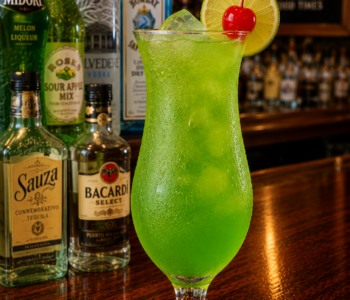 A midori madness cocktail recipe served in a hurricane glass with lime and cherry garnish on a neighborhood bar counter.