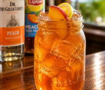 A peach iced tea cocktail recipe served in a mason jar with peach slices, lemon garnish, and ice on a wooden bar counter.