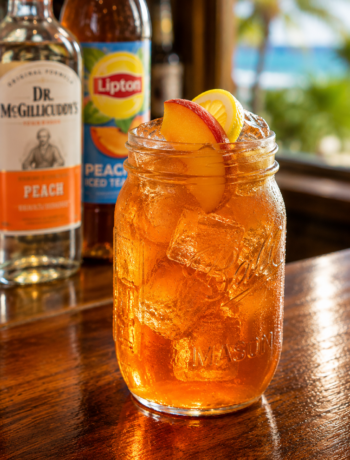 A peach iced tea cocktail recipe served in a mason jar with peach slices, lemon garnish, and ice on a wooden bar counter.