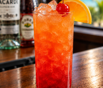 A red rum cocktail recipe served in a highball glass with crushed ice, orange slice, and cherry garnish on a wooden bar counter.